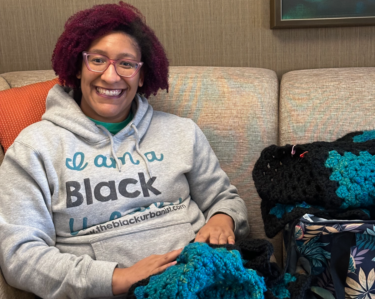 Kristen smiling, with fuchsia kinky curly hair and cat eye glasses, wearing an I am a Black Urbanist hoodie and holding a crocheted sweater she's making, is seated on a couch in a Hyatt Place hotel room