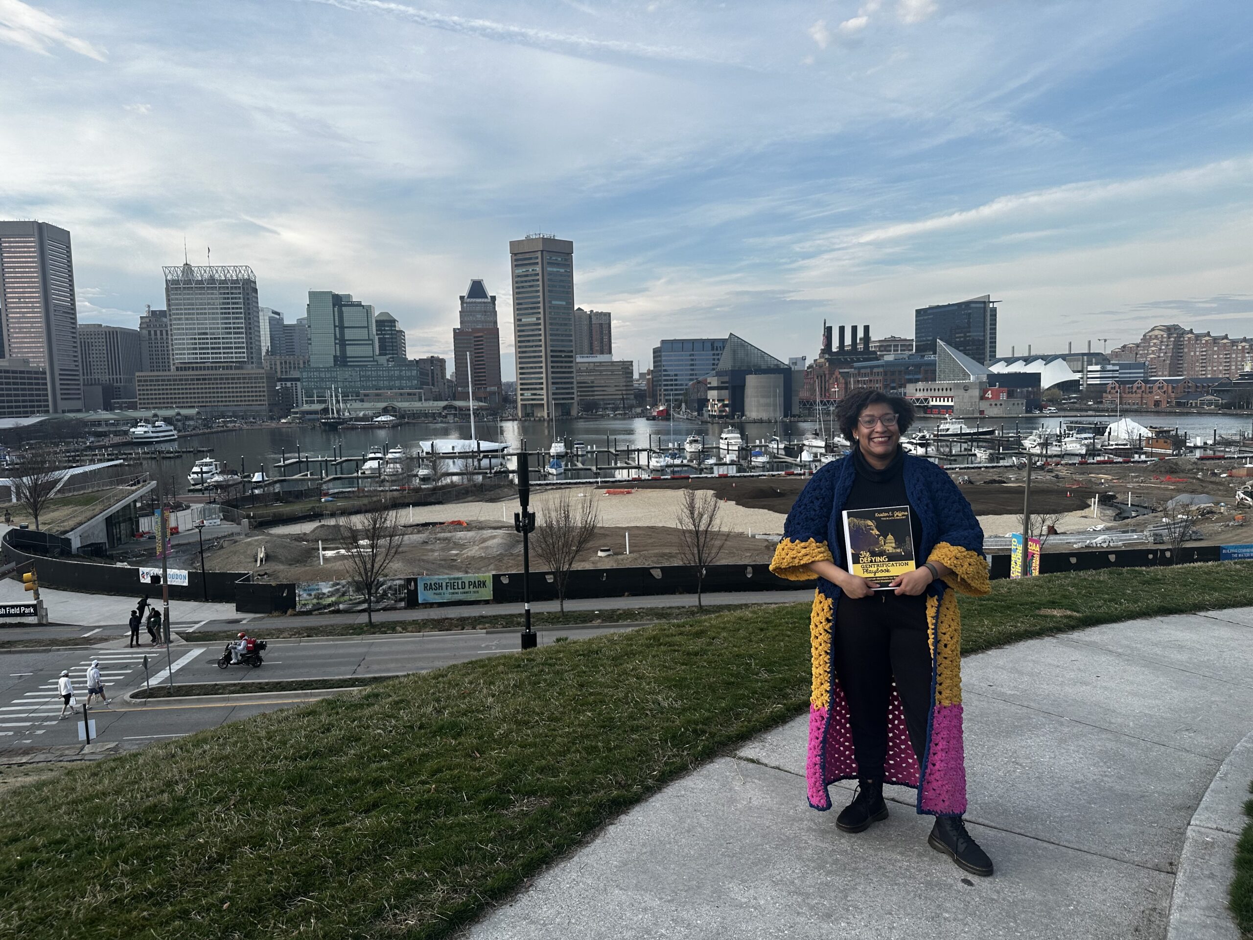 Kristen standing facing the camera on Federal Hill with the Baltimore Maryland skyline behind them. They are wearing a navy, yellow, and pink striped coat they crocheted and holding their book The Defying Gentrification Playbook